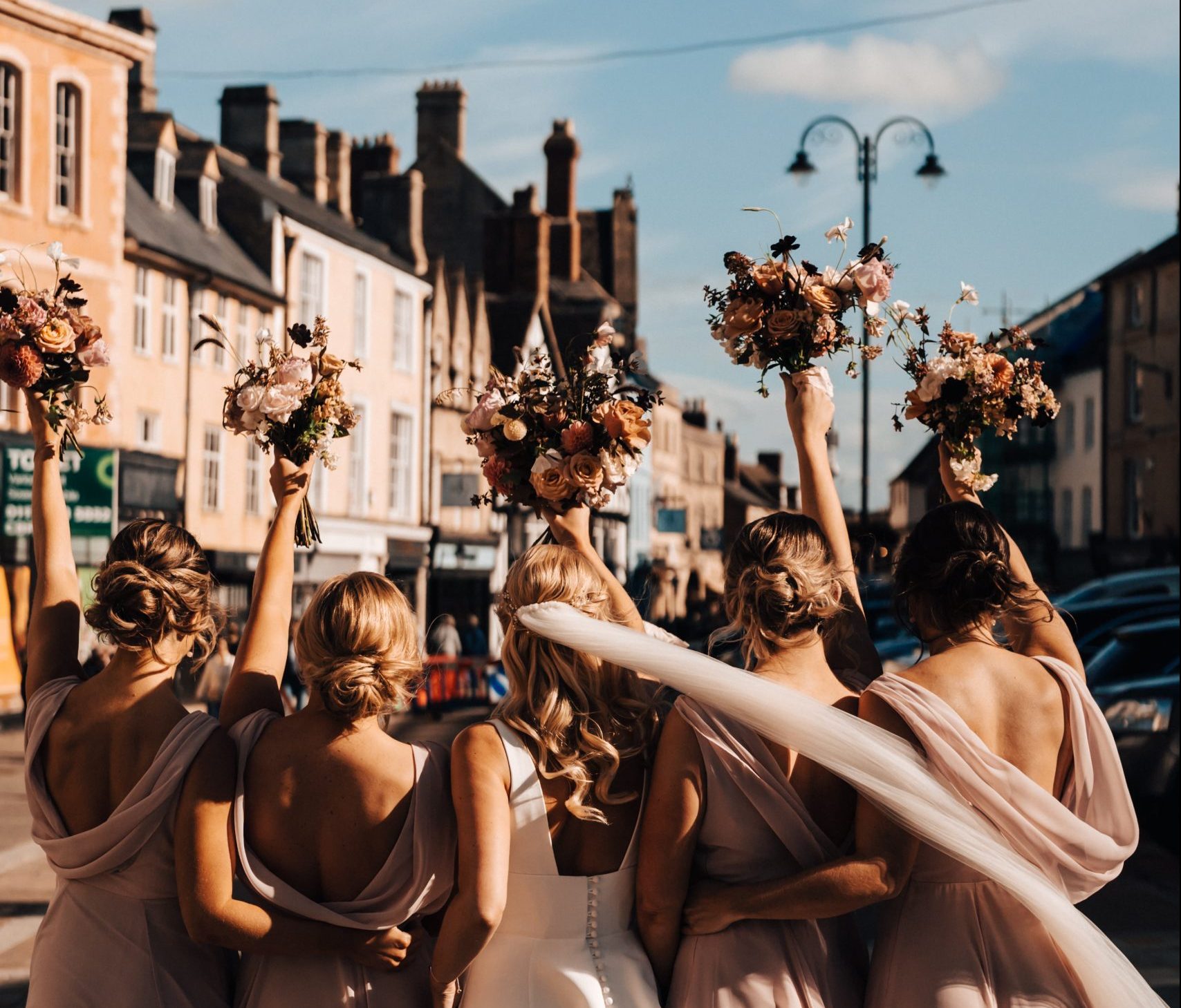 Bride and 4 bridesmaids with their back to the camera with arms extended holding bridal bouquets on a street in Cirencester